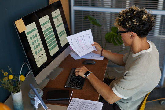 Young adult Caucasian man sitting at desk developing mobile app interface, holding wireframe sketches in one hand while working on computer with smartphone screen designs displayed