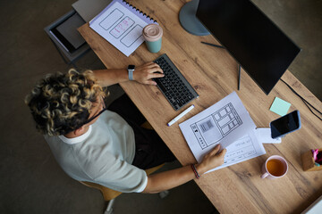 Young adult Caucasian man working at desk holding website wireframe sketch while typing on...