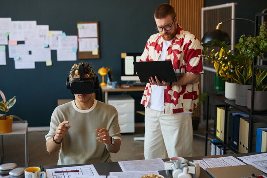 Young adult Caucasian man wearing virtual reality headset interacting with digital interface while standing bearded Caucasian man holding tablet observing in modern office workspace - Powered by Adobe