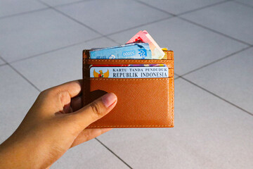 A hand holding a wallet with money (100k and 50k rupiah) and ID (KTP).