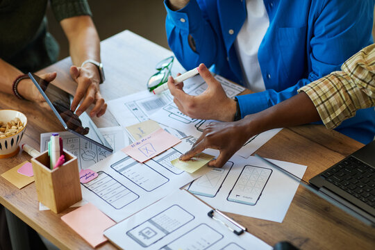 Diverse group of young adult men and women collaborating on mobile app interface design, using digital tablet and paper wireframes, discussing project details at shared workspace