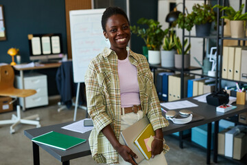 Portrait of young adult Black woman smiling while standing in modern office holding folders and...