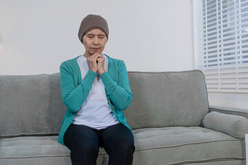 An elderly Asian woman wearing a headwrap sits quietly on a sofa, lost in deep thought and...
