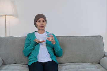 An elderly woman with cancer sits on a sofa at home, wearing a headwrap and drinking water. She...