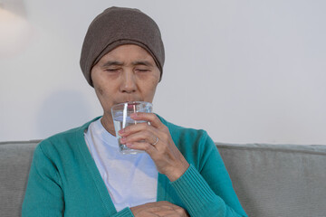 An elderly woman with cancer sits on a sofa at home, wearing a headwrap and drinking water. She...