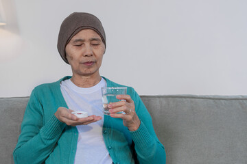 An elderly woman with cancer sits on a sofa at home, wearing a headwrap and drinking water. She...