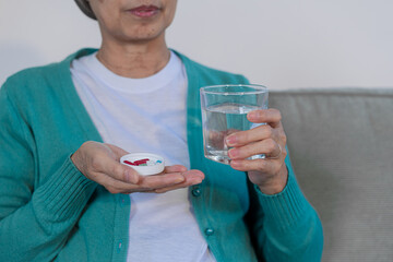 An elderly woman with cancer sits on a sofa at home, wearing a headwrap and drinking water. She...
