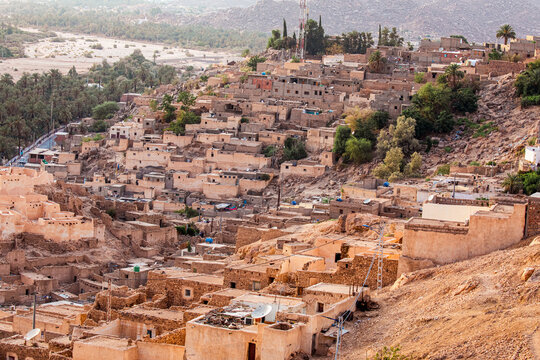A sweeping high-angle view captures the unique character of Djanet, a vital oasis town nestled within Algeria's vast Sahara Desert. The settlement is composed of traditional, earthy-toned buildings cl