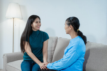 An Asian mother and daughter sit closely on a sofa, sharing a warm embrace. Their gentle...