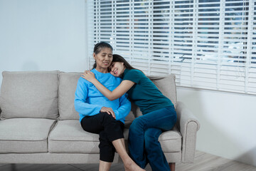 An Asian mother and daughter sit closely on a sofa, sharing a warm embrace. Their gentle...