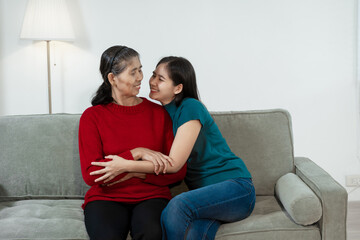 An Asian mother and daughter sit closely on a sofa, sharing a warm embrace. Their gentle...