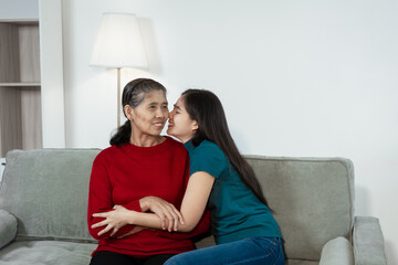 An Asian mother and daughter sit closely on a sofa, sharing a warm embrace. Their gentle...