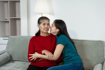 An Asian mother and daughter sit closely on a sofa, sharing a warm embrace. Their gentle...