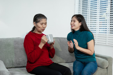 An Asian mother and daughter sit closely on a sofa, sharing a warm embrace. Their gentle...