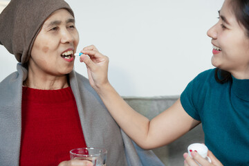 An Asian young woman carefully cares for her ill mother at home, sitting on the sofa, offering...