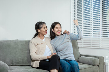 An Asian daughter gently holds her elderly mother’s hand as they sit on a sofa at home, sharing a...