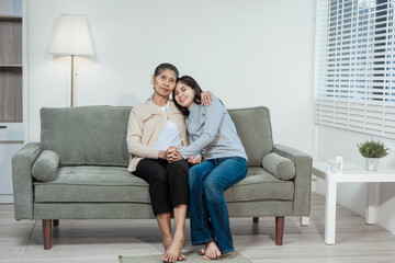 An Asian daughter gently holds her elderly mother’s hand as they sit on a sofa at home, sharing a...