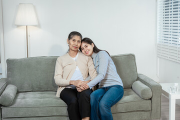 An Asian daughter gently holds her elderly mother’s hand as they sit on a sofa at home, sharing a...