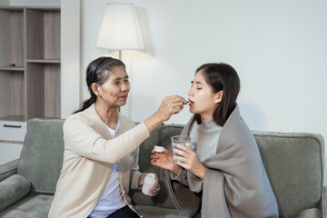 An elderly Asian woman gently checks her sick daughter's forehead, offering care and comfort. The young woman,wrapped in a blanket, holds a glass of water while being looked after in the living room