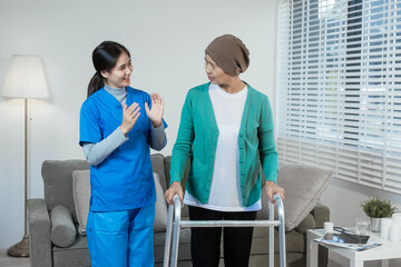 A young Asian nurse assists an elderly female cancer patient, wearing a headscarf, with using a...