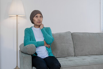 An elderly Asian woman wearing a headwrap sits quietly on a sofa, lost in deep thought and...