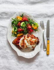 Chicken schnitzel with tomato sauce and mozzarella and fresh vegetable salad on a light background, top view