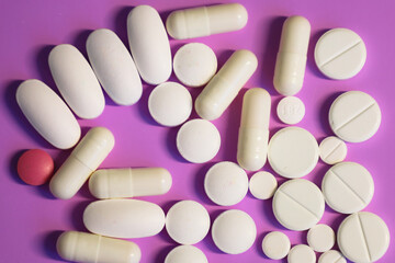 A group of white tablets and capsules, with one pink pill, scattered on a bright purple background,...