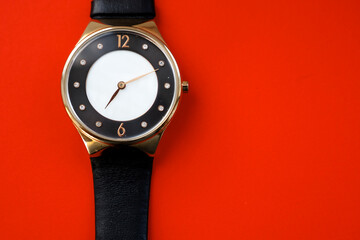A round golden wristwatch with a black leather strap lies on a bright red background. Its classic...