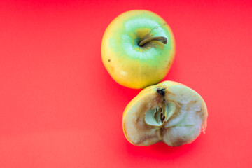 A whole and a cut rotten apple lie on a solid red background. The image illustrates the process of decay, highlighting the contrast between the external freshness and internal damage.