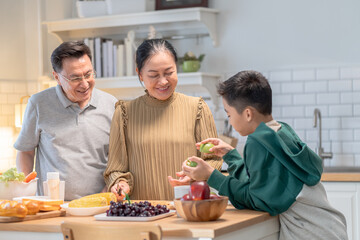 Joyful Asian grandparent, bonding at home lifestyle, warm moment of elderly couple enjoying coffee conversation while grandson stands together, cozy holiday kitchen with relaxed background atmosphere