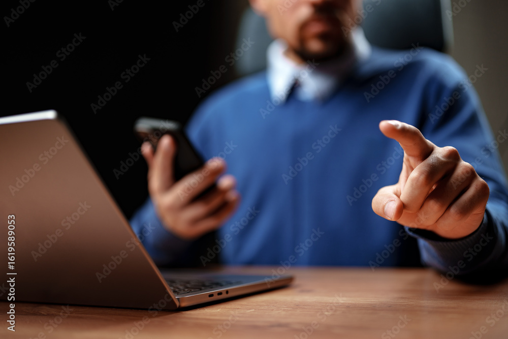 Poster engaged professional communicating via smartphone while using laptop in a modern workspace environme