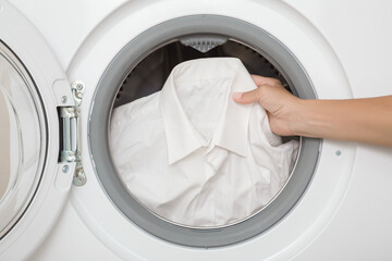 Young adult woman hand holding classic male shirt in opened washing machine. White clothes care. Closeup. Front view.
