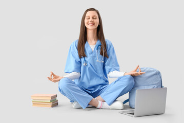 Female medical intern with books and laptop meditating on light background