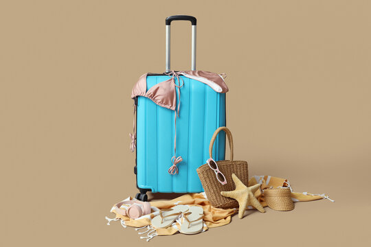 Blue suitcase with stylish swimsuit, wicker bag, flip flops and modern headphones on brown background