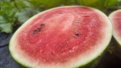 Close-up of a freshly cut watermelon showing juicy red flesh and seeds, placed on the ground in a watermelon field surrounded by green leaves. Represents natural agriculture and fresh tropical fruit h
