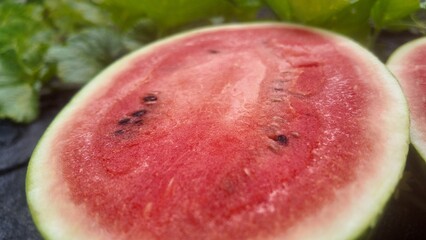 Close-up of a freshly cut watermelon showing juicy red flesh and seeds, placed on the ground in a watermelon field surrounded by green leaves. Represents natural agriculture and fresh tropical fruit h