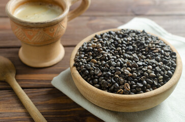 Green coffee beans in a wooden bowl with a ceramic coffee cup on a brown table. Alternative drink concept. Rustic style. Selective focus. Horizontal orientation.