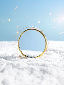 A minimalist and elegant studio shot of a golden circular ring with delicate fairy lights wrapped around it, standing upright in a bed of pristine white snow against a clear blue background with subtl