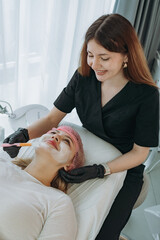 professional aesthetician applies a creamy face mask on young woman face using a brush. Doctor looking at camera