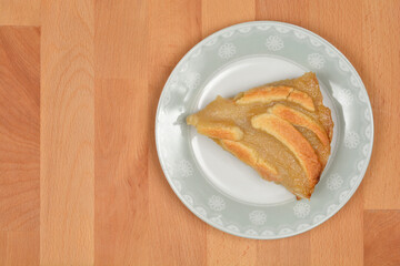 A triangular slice of apple pie with baked crust strips rests on a light blue plate with a decorative rim, positioned on a light wooden surface