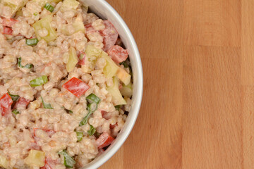 A portion of a vibrant barley salad with peppers, tomatoes, and green onions mixed with mayonnaise is seen in a white bowl, partially placed on a light wooden surface