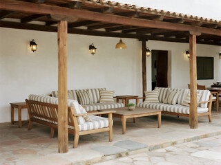A rustic, wooden outdoor seating area with striped cushions, surrounded by white walls and a clay-tiled roof, illuminated by warm pendant lights.