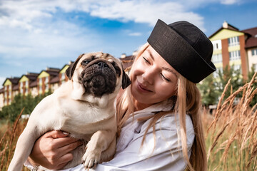 Young happy beautiful woman holding and hugging her beloved dog during a walk with harness and black leash in a city park. Street photography. Favorite pets.