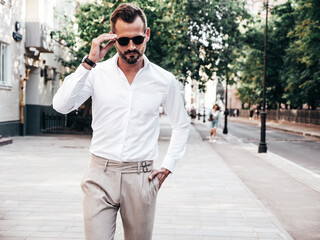 Portrait of handsome confident stylish hipster lambersexual model.Sexy modern man dressed in white shirt and trousers. Fashion male posing on street background in Europe city at sunset. In sunglasses