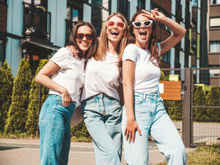 Three young beautiful smiling hipster female in trendy summer same clothes. Sexy carefree women posing on the street background.Positive models having fun in sunglasses.Hugging. Cheerful and happy