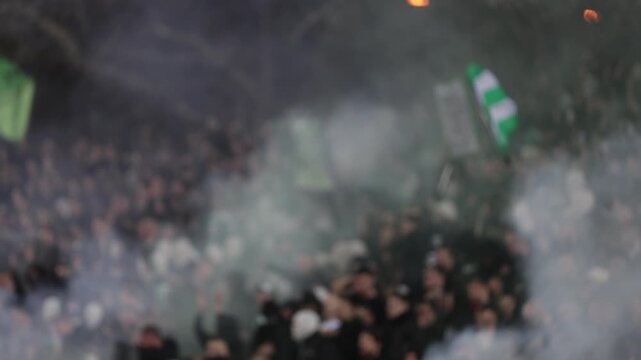 Many fans supporting soccer team at stadium. Blurred crowd of people at football stadium. Torch lights and smoke bombs