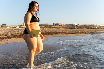 Curvy woman walking on the beach holding a book at sunset