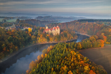 Czocha Castle aerial shot in autumn, surrounded by colorful trees, nestled by a lake, medieval architecture with warm seasonal tones, located in southwestern Poland.