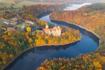 Czocha Castle aerial shot in autumn, surrounded by colorful trees, nestled by a lake, medieval architecture with warm seasonal tones, located in southwestern Poland.