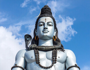 Lord Shiva sculpture under clear blue sky with calm expression, divine snake adornment and traditional jewelry in sacred pose